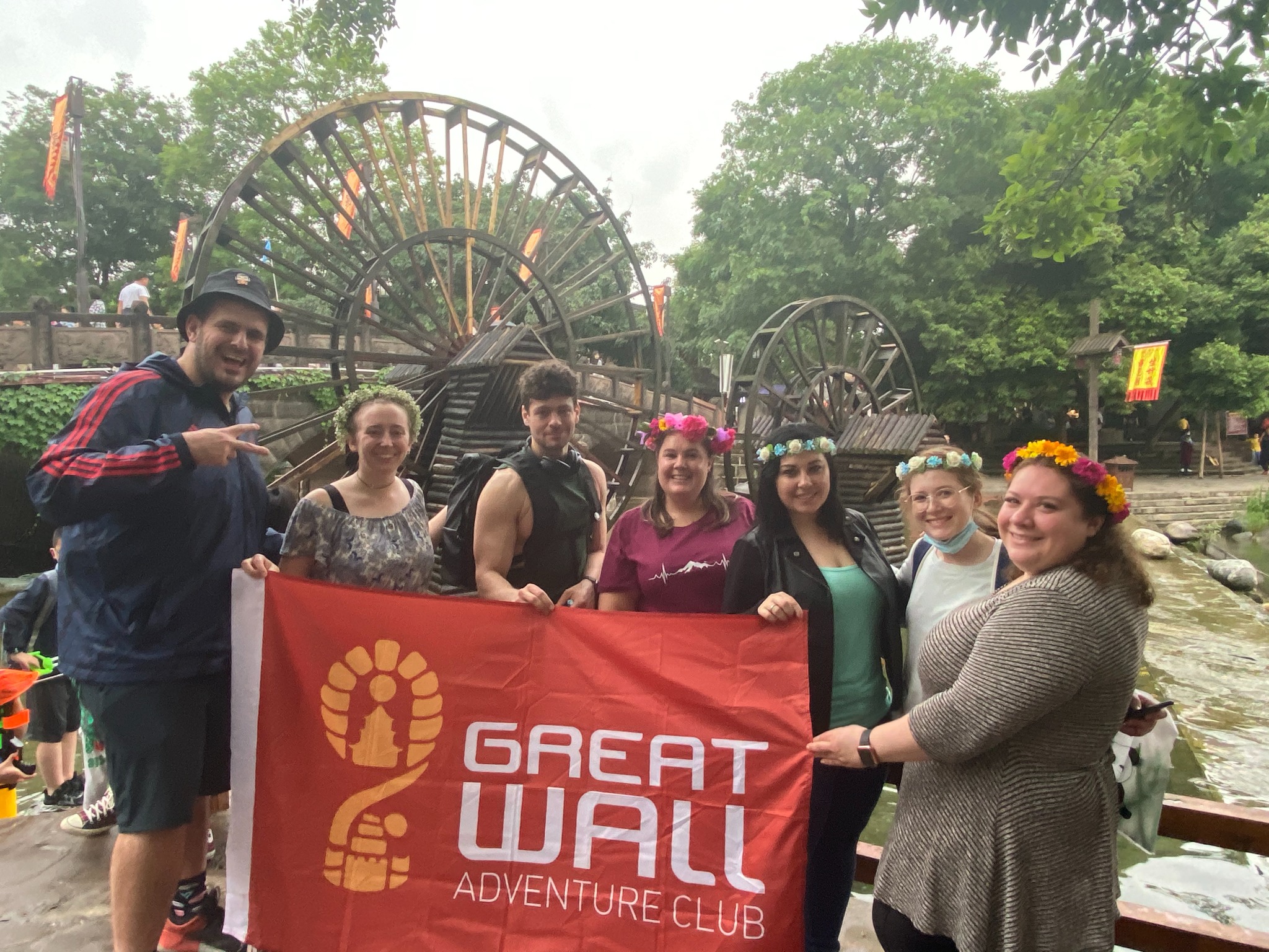 Traveler group holding a Great Wall Adventure Club banner near a wooden waterwheel