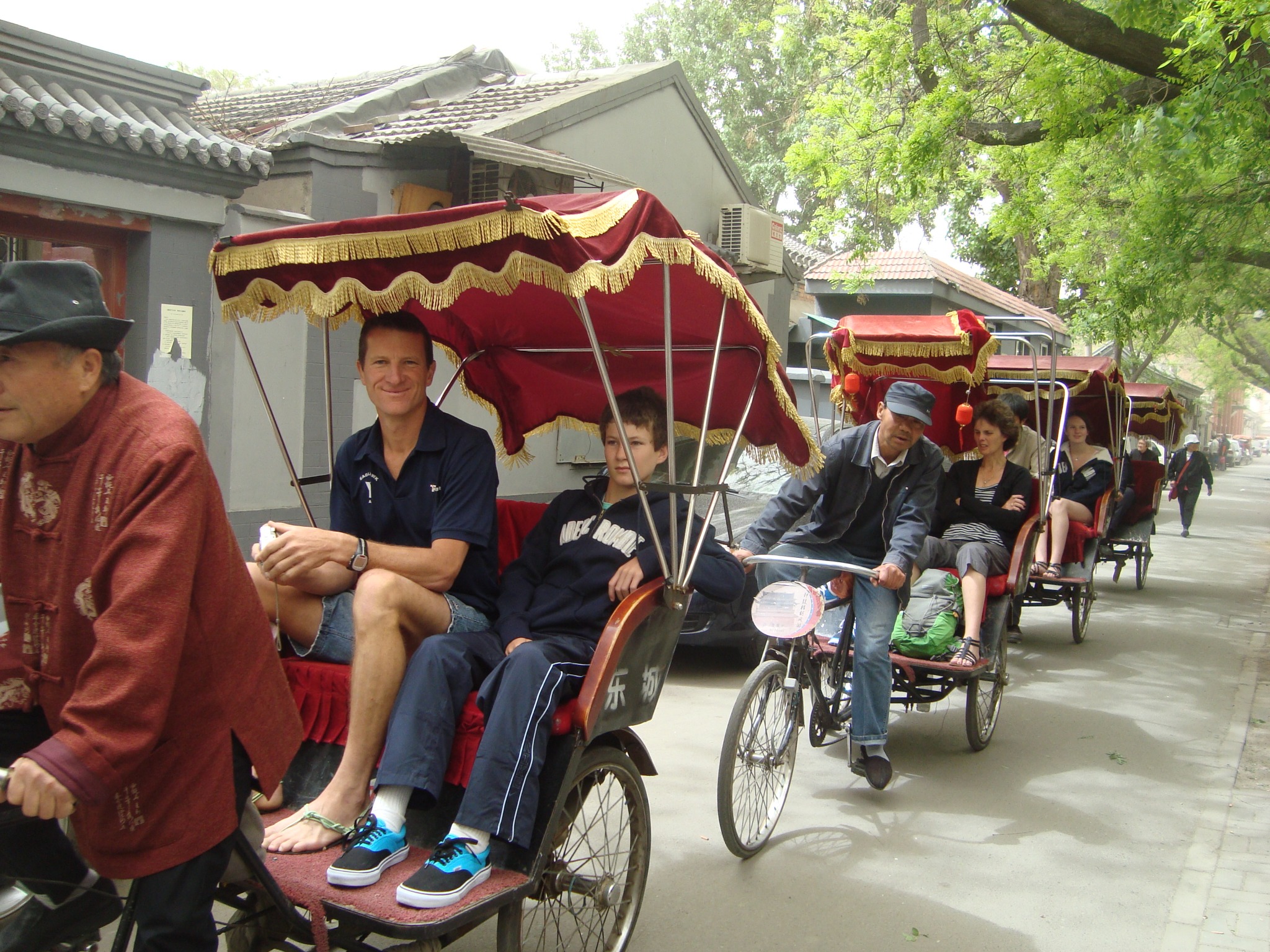 Travelers riding red-canopied rickshaws through a tree-lined Beijing hutong